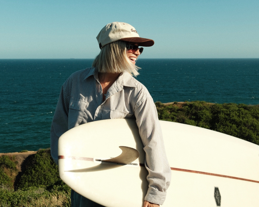 Person holding a surfboard with ocean in the background wearing a brown vintage baseball cap