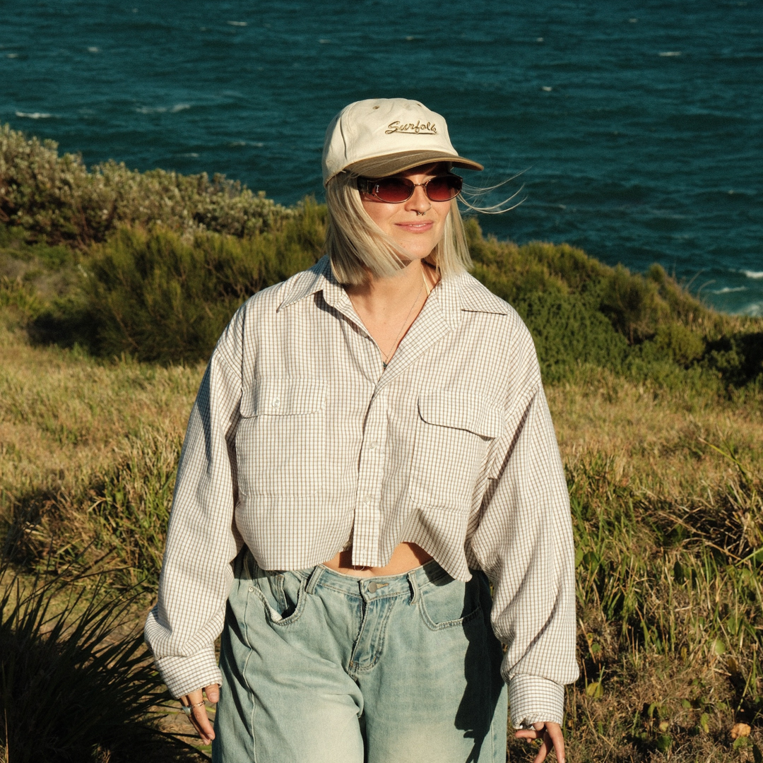 Person wearing a cap and sunglasses standing on a grassy hill with ocean in the background