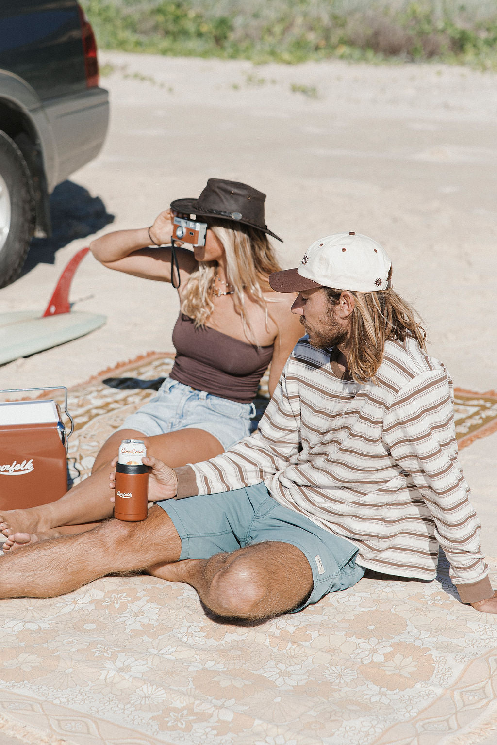 Two people sitting on a blanket outdoors with a cooler and drinks.