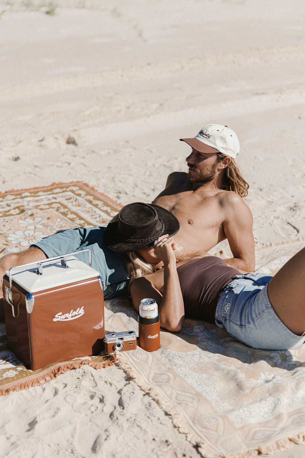 A man and a woman relax on a patterned beach blanket on a sandy beach. A brown Scabbly cooler, a brown bottle, and a small camera are placed beside them.