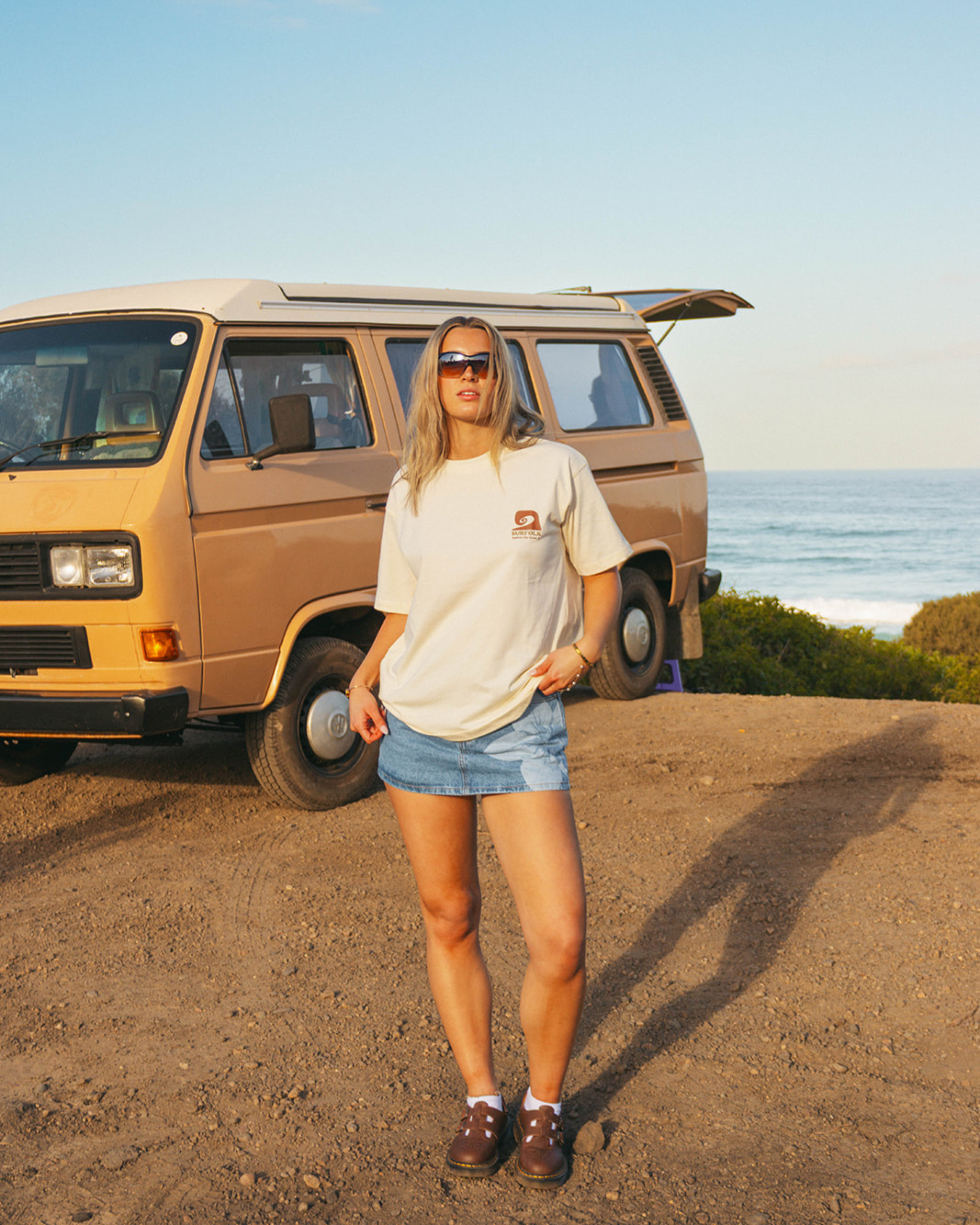 A woman stands outdoors in front of a tan vintage van and the ocean, wearing a cream graphic T-shirt, denim shorts, and brown shoes. The product appears to be the T-shirt, shown in a real environment with a person present, so subject_type is Lifestyle.