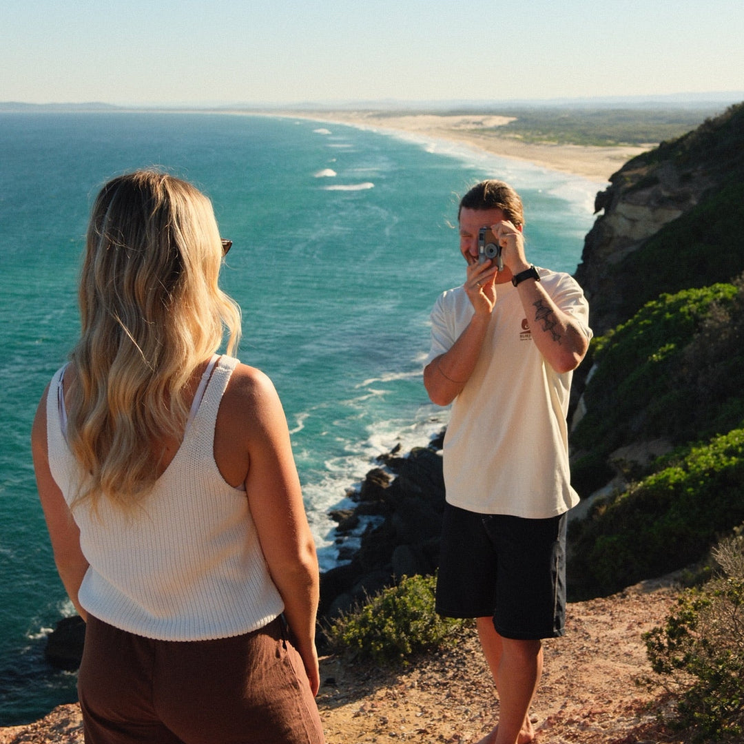 A woman in a white tank top and brown pants stands on a cliffside overlooking the ocean, while a man in a white t-shirt and black shorts takes a photo of the scenic coastline.