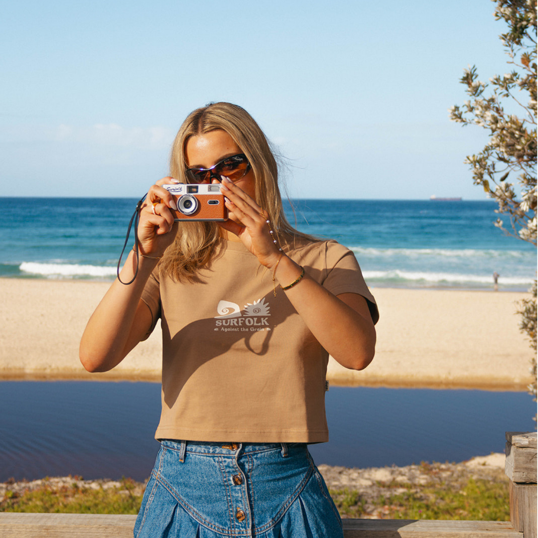 A woman wearing a tan cropped t-shirt with a 'SURFOLK' graphic and blue denim skirt, holding a vintage camera in front of her face, with a beach and ocean in the background.