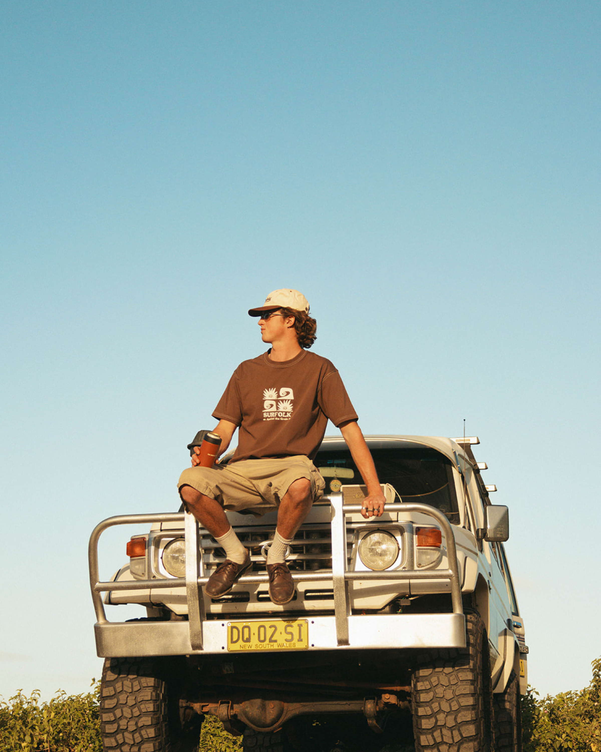 A man wearing a brown graphic t-shirt, beige shorts, and a white cap sits on the front bumper of a silver off-road vehicle under a clear blue sky.