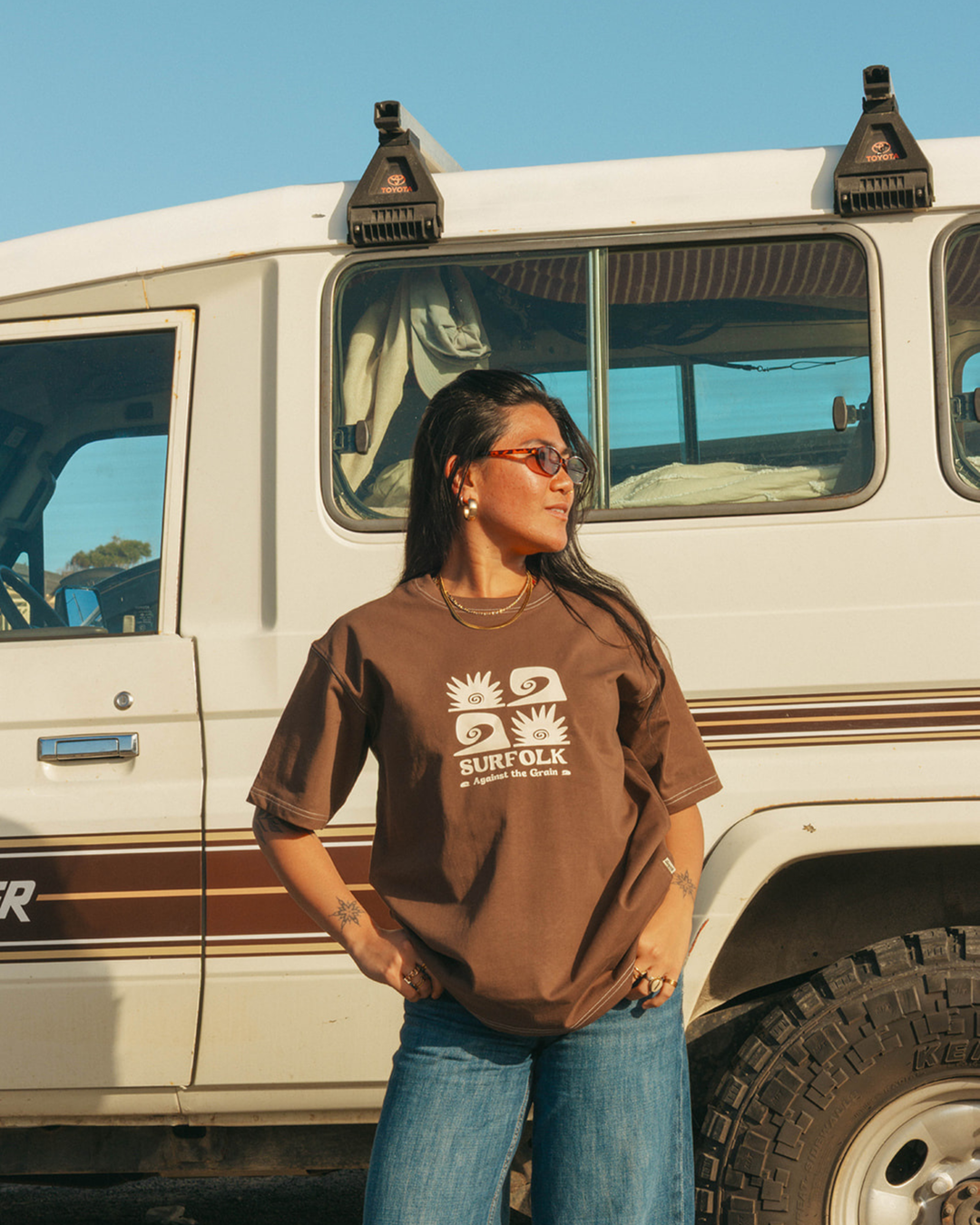 A woman wearing a brown graphic t-shirt and blue jeans stands in front of a white vintage vehicle under a clear blue sky.