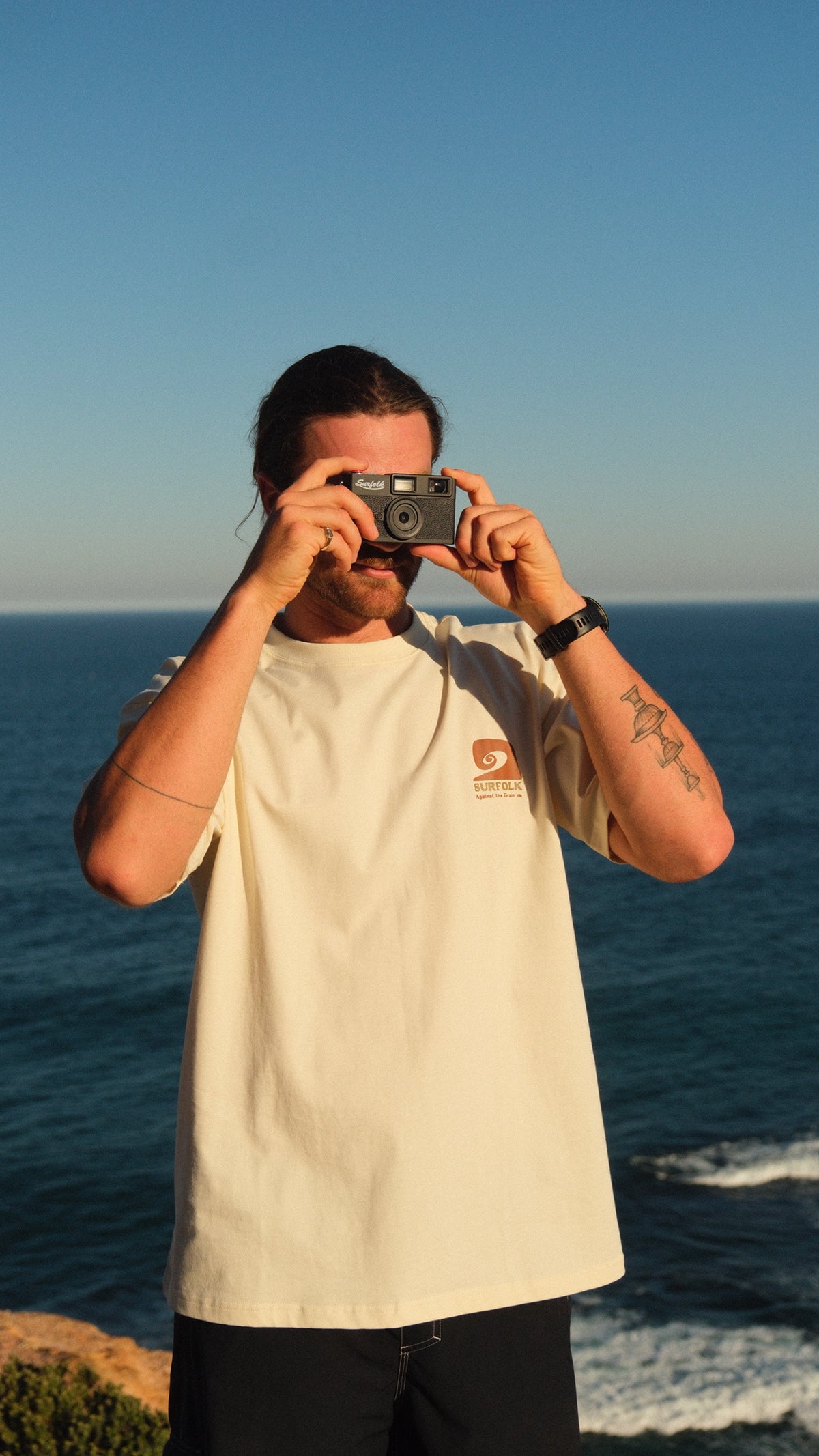 A man wearing a cream-colored t-shirt with a small orange logo on the left chest stands on a cliff overlooking the ocean, holding a vintage camera up to his face. The background features a clear blue sky and sea.