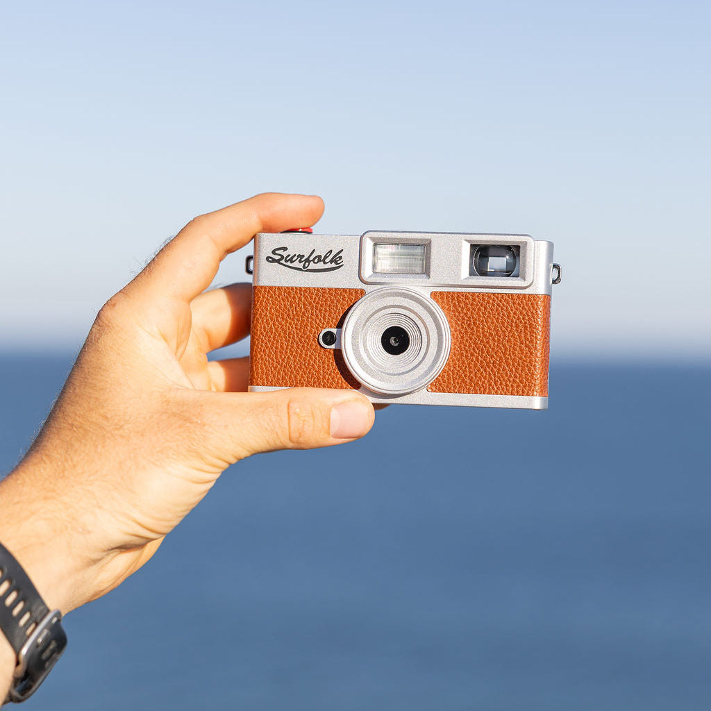 A hand holding a small Zennin camera with a silver body and textured brown leatherette front, set against a blurred blue ocean and sky background.
