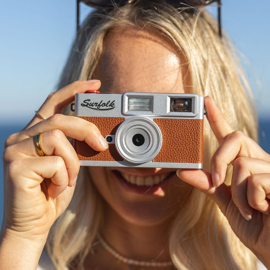 A woman with blonde hair holding a Surfolk vintage-style retro digital camera in front of her face, with sunglasses on her head and a blue sky background.