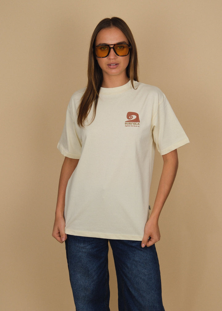 A woman modeling an oversized cream-colored t-shirt with a small brown logo on the left chest, paired with dark blue jeans, set against a solid beige background.