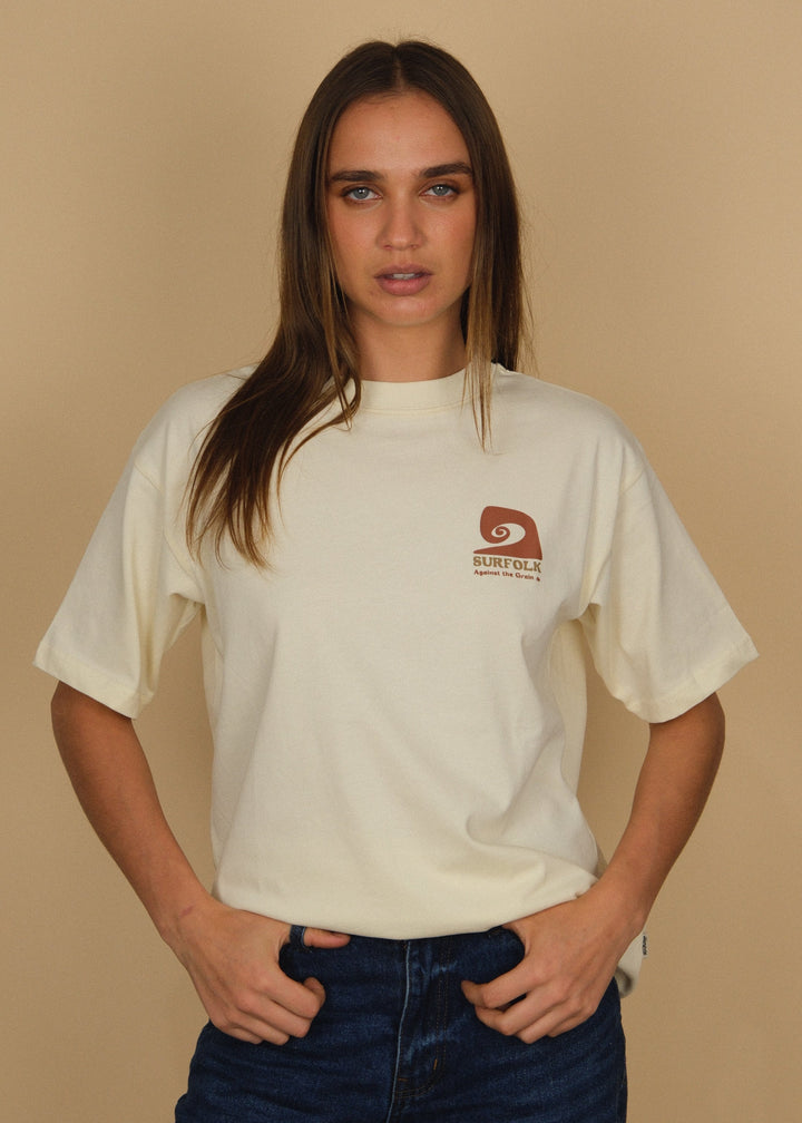 A woman modeling a cream-colored short-sleeve t-shirt with a small brown logo on the left chest, paired with dark blue jeans, against a solid beige background.