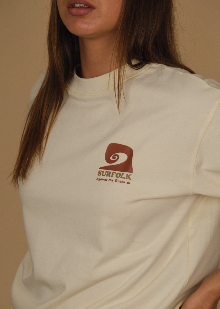 A woman wearing a cream-colored crewneck t-shirt with a brown Surfcloud logo on the left chest, paired with dark blue jeans, set against a plain beige background.