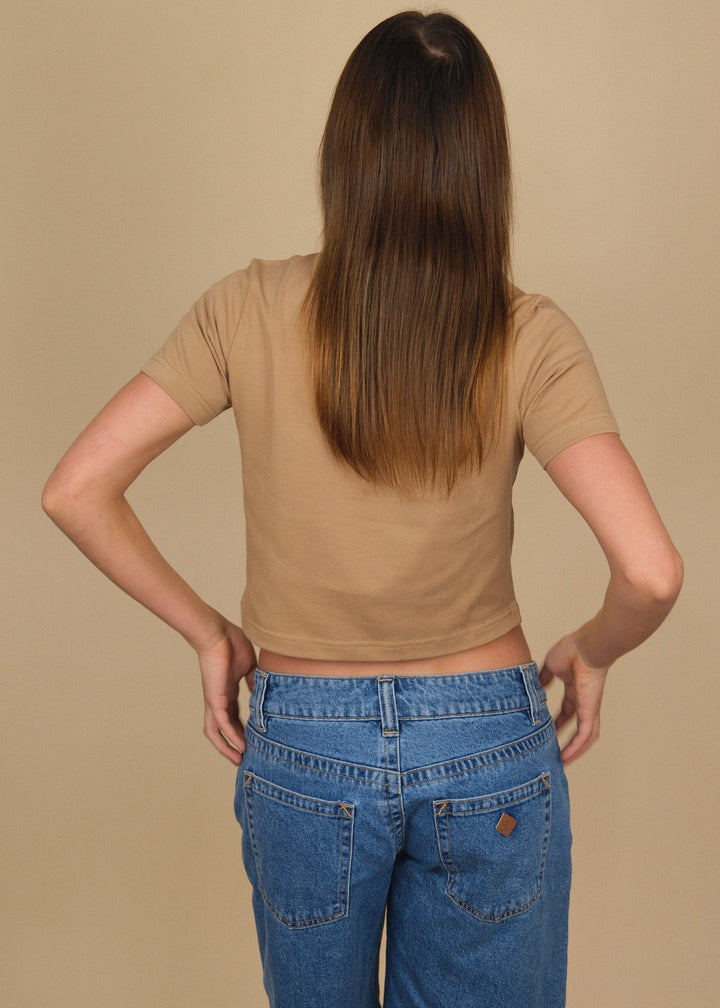 A woman viewed from behind wearing a tan short-sleeve crop top and blue denim shorts against a solid beige studio background.