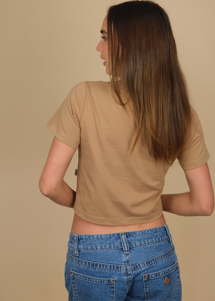 A woman with long brown hair is shown from the back wearing a beige short-sleeve crop top and blue jeans against a solid beige background.