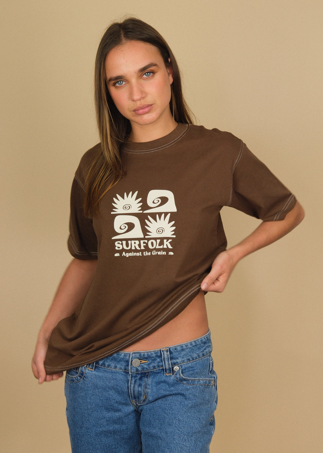 A woman modeling a brown short-sleeve t-shirt with a white graphic print that reads 'SURFOLK' and 'Against All Crops', paired with blue denim shorts, against a solid beige background.