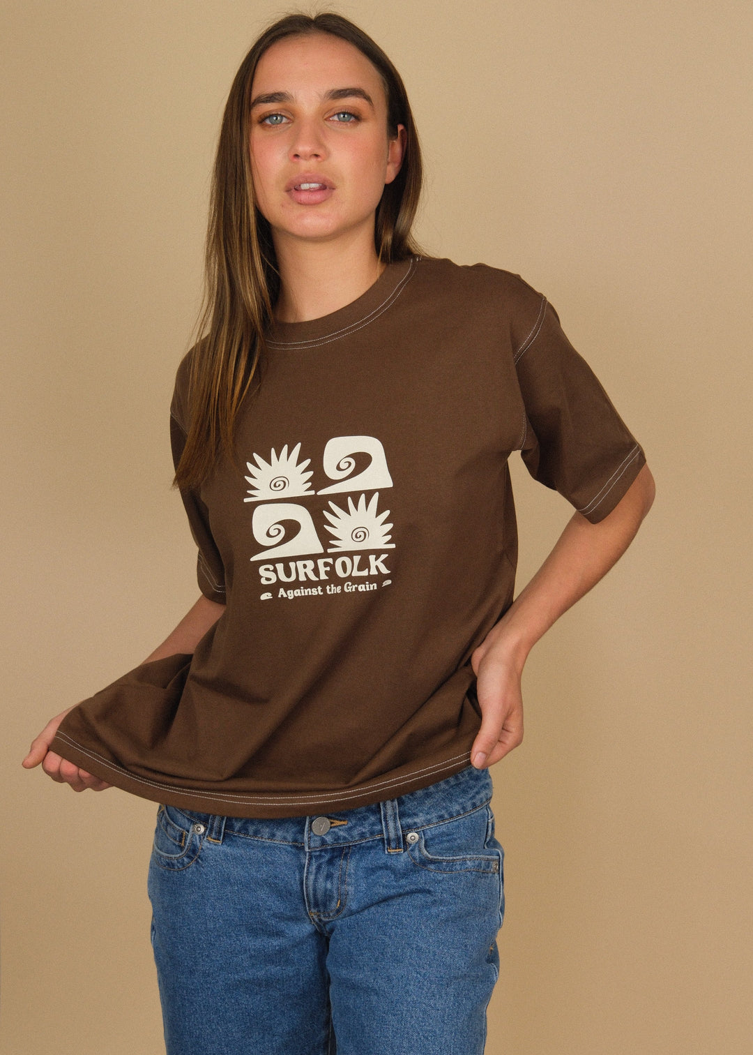 A woman modeling a brown t-shirt with a white surf-themed graphic that reads &