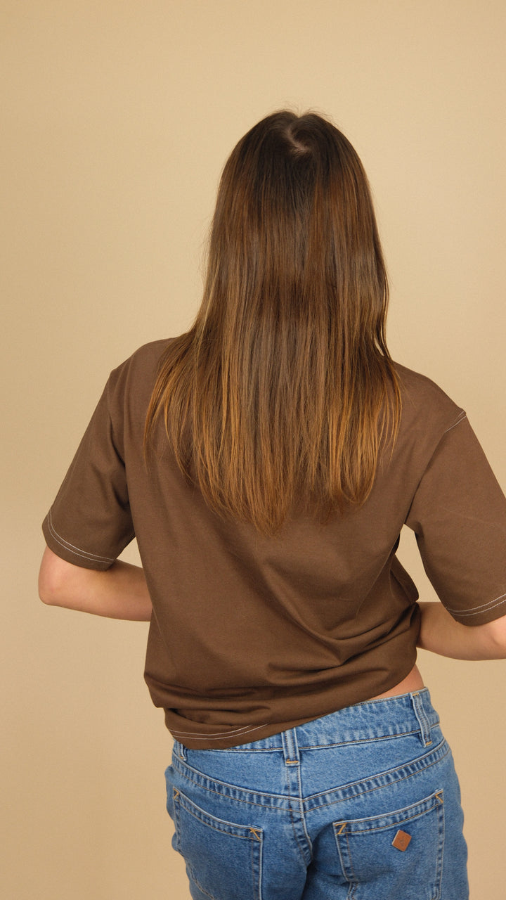 A person viewed from behind wearing a brown short-sleeve t-shirt and blue jeans, standing against a solid beige background.