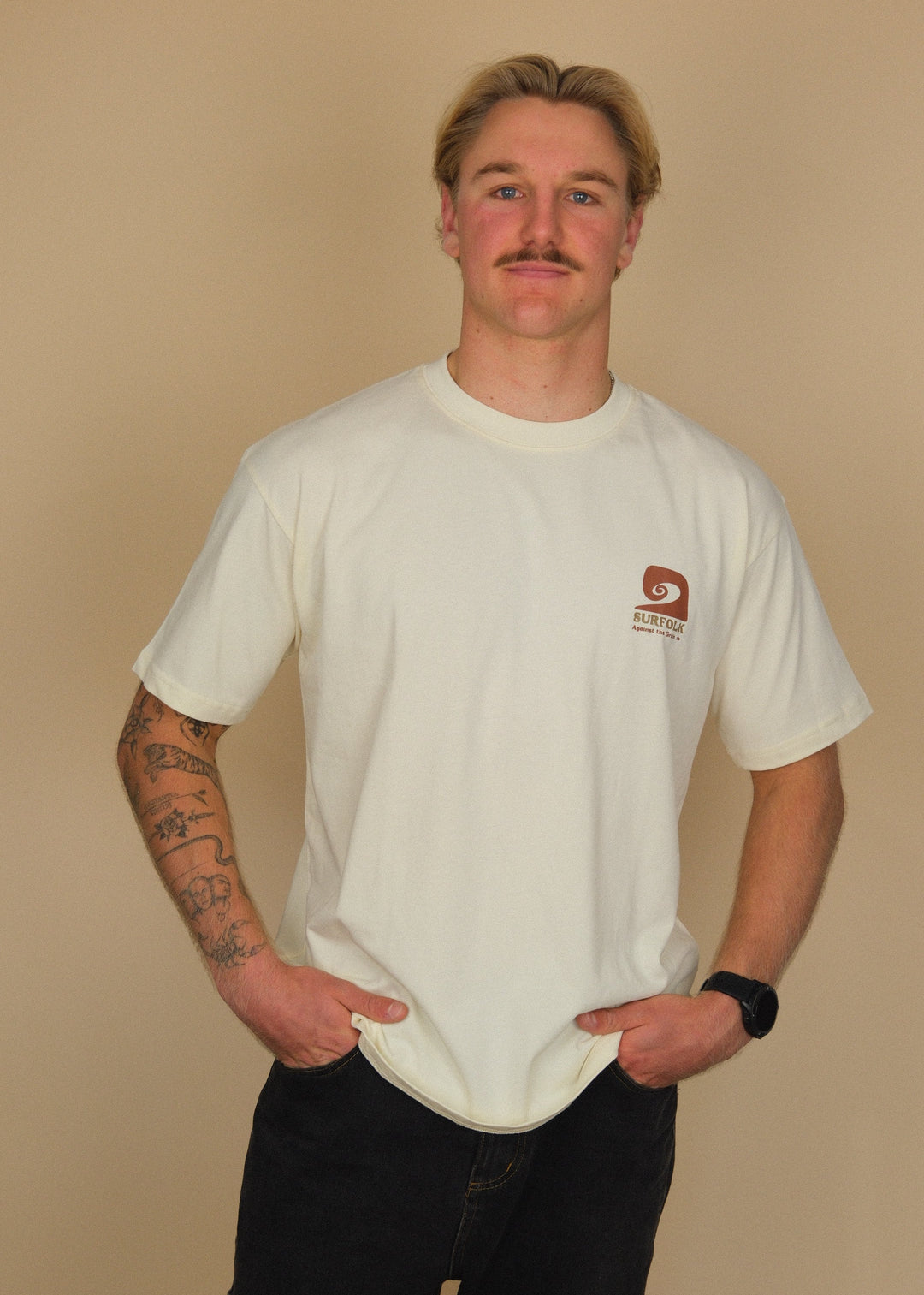 A man with a mustache modeling a cream-colored crewneck t-shirt with a small brown logo on the left chest, paired with black shorts, standing against a solid beige background.