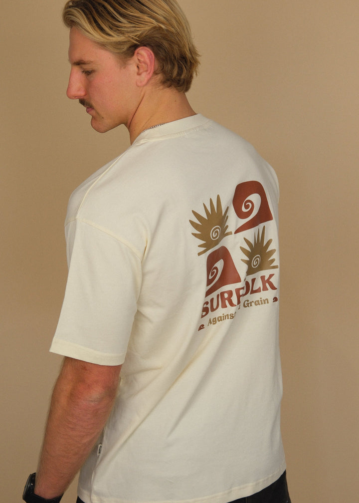 A man wearing a cream-colored t-shirt with a large brown and red Surplk logo printed on the back, shown from a side-back angle against a solid beige background.