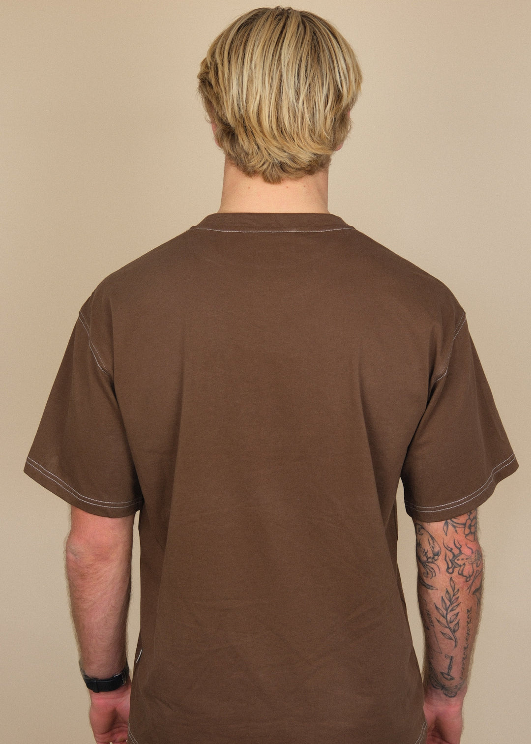 A man viewed from behind wearing a brown short-sleeve t-shirt and dark jeans, standing against a plain beige background.
