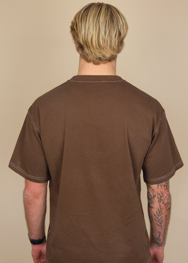 A man viewed from behind wearing a brown short-sleeve t-shirt and dark jeans, standing against a plain beige background.