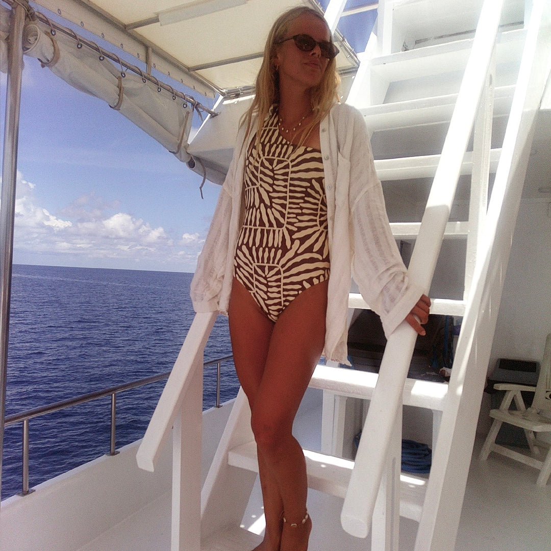 A woman wearing a brown and beige zebra-print one-piece swimsuit, an open white linen cover-up, and sunglasses stands on the white wooden stairs of a boat with the ocean in the background.
