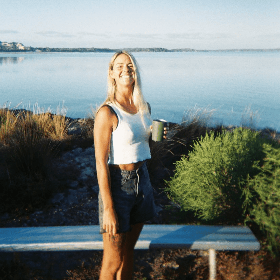 A 35mm film image of a Woman holding a mug by a lake with a bench in the foreground.
