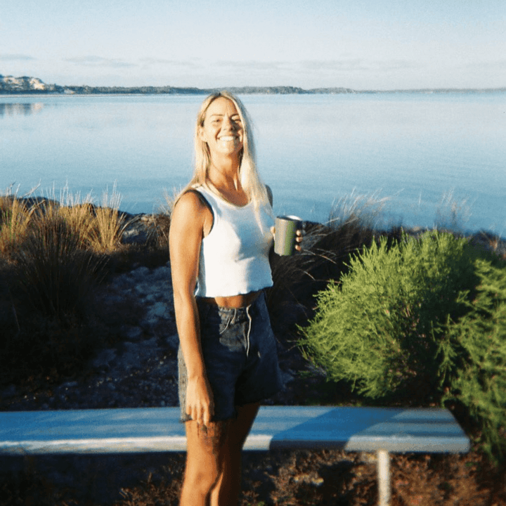 A 35mm film image of a Woman holding a mug by a lake with a bench in the foreground.