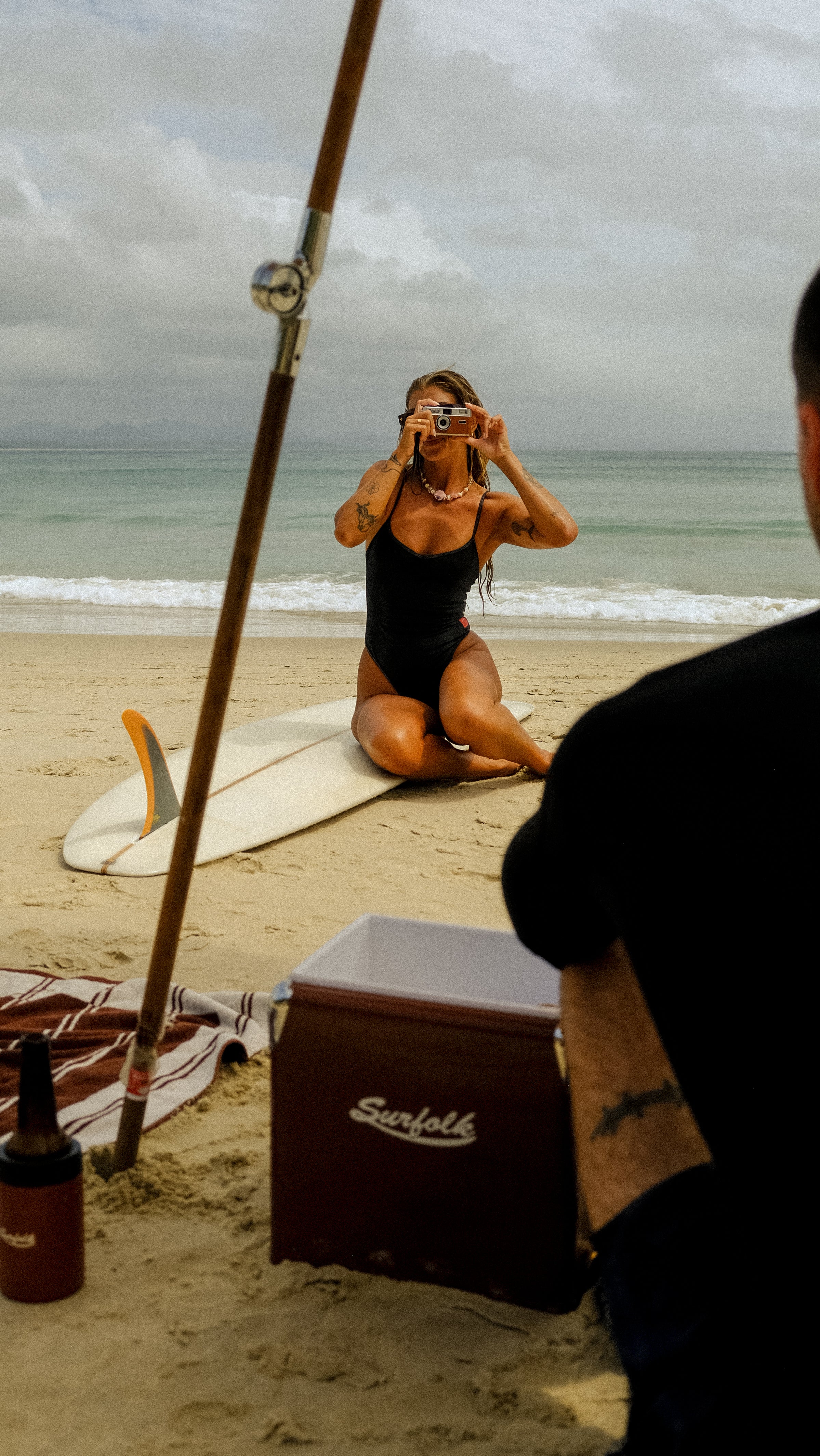 Woman in a black swimsuit taking a photo on a beach with a cloudy sky.